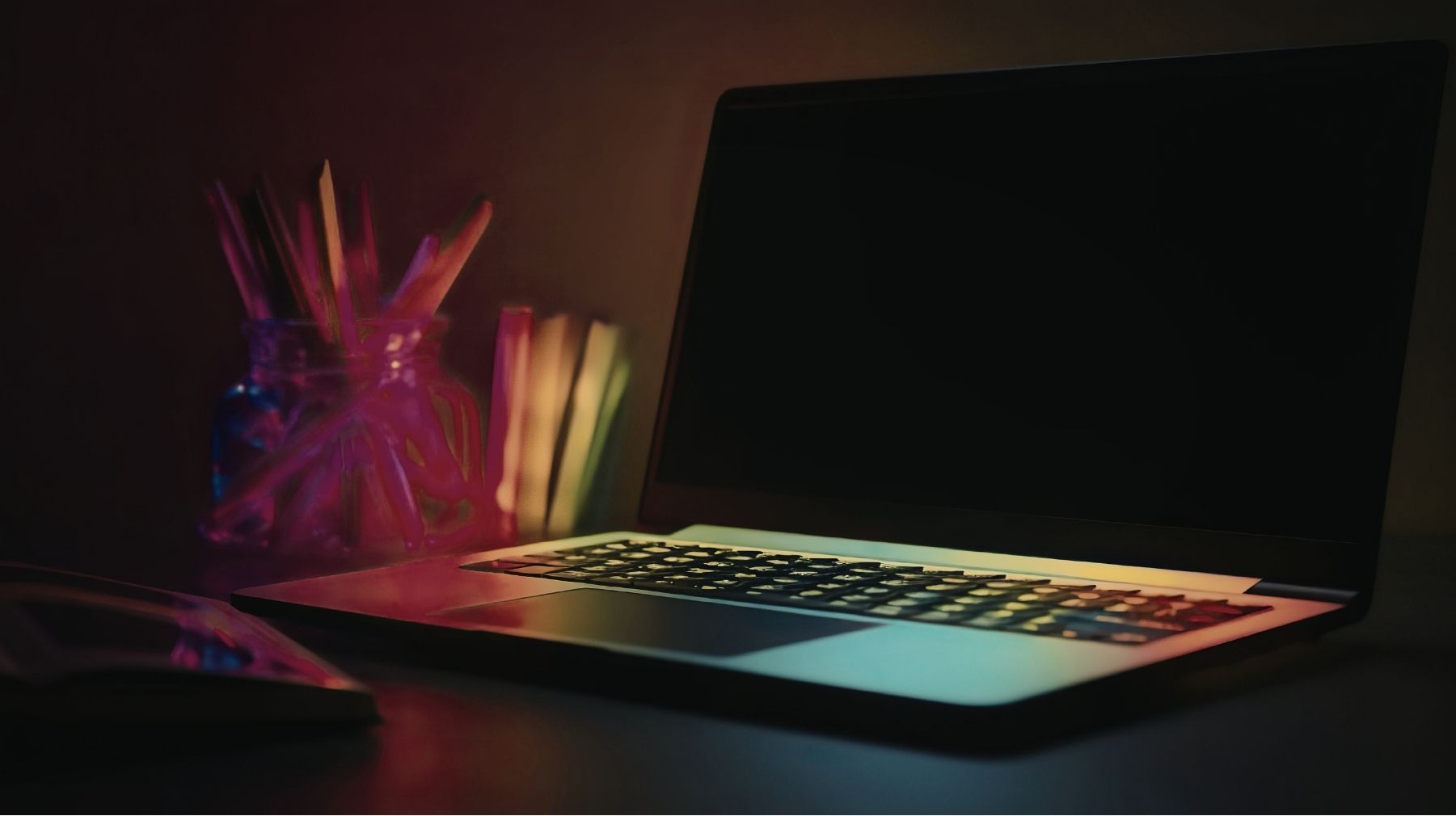 A laptop with a blank screen sits on a desk next to a glass jar filled with pens and pencils, and a few books, under dim, colorful lighting.
